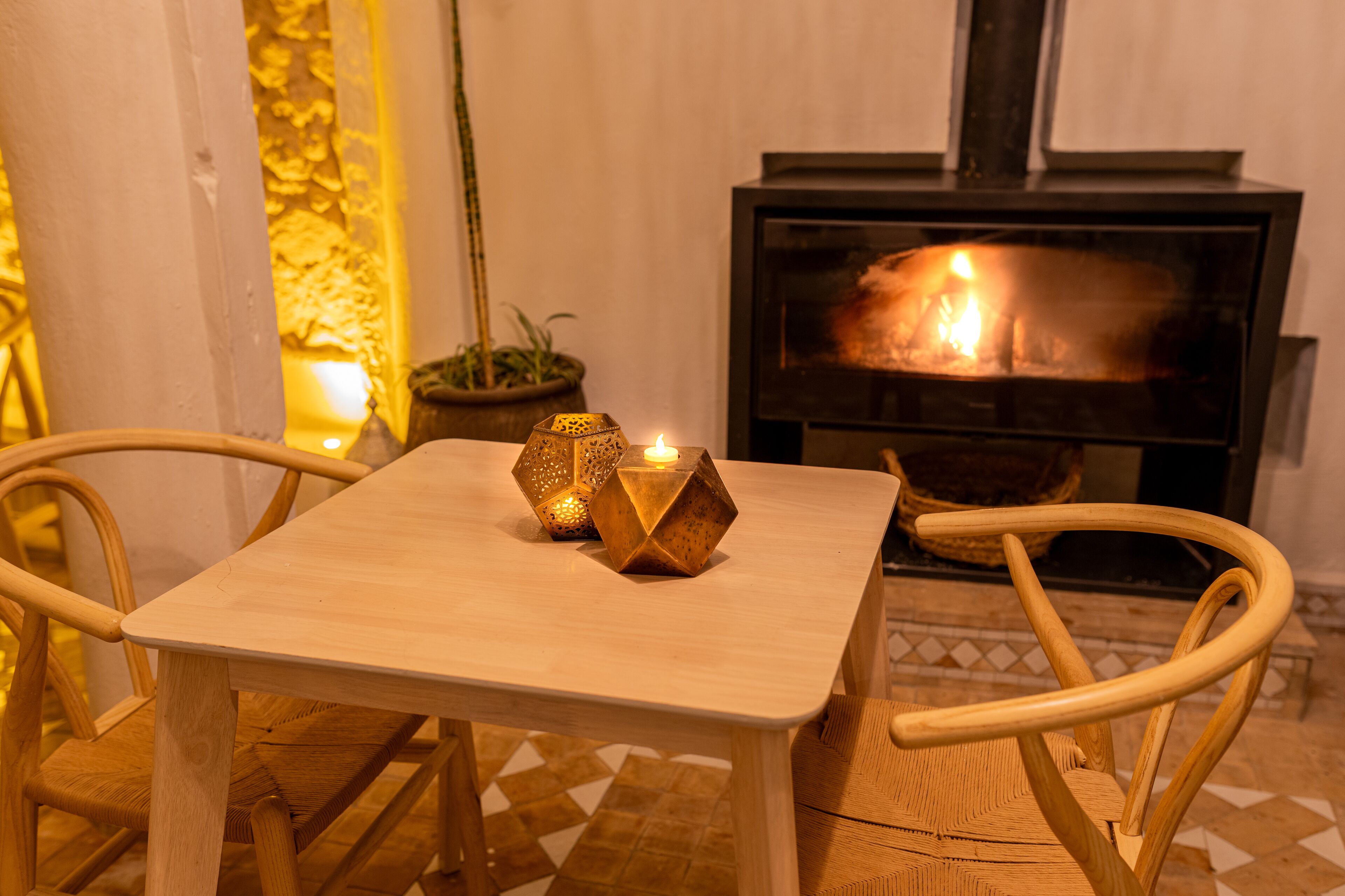 A wooden table with two chairs, a lit candle, and a fireplace in the background.