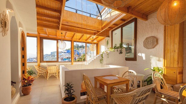A spacious room with a wooden ceiling, large windows offering a view of the city, wicker furniture, and potted plants.