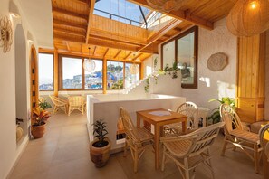 A spacious room with a wooden ceiling, large windows offering a view of the city, wicker furniture, and potted plants. - RIAD NILA  (Chefchaouen)
