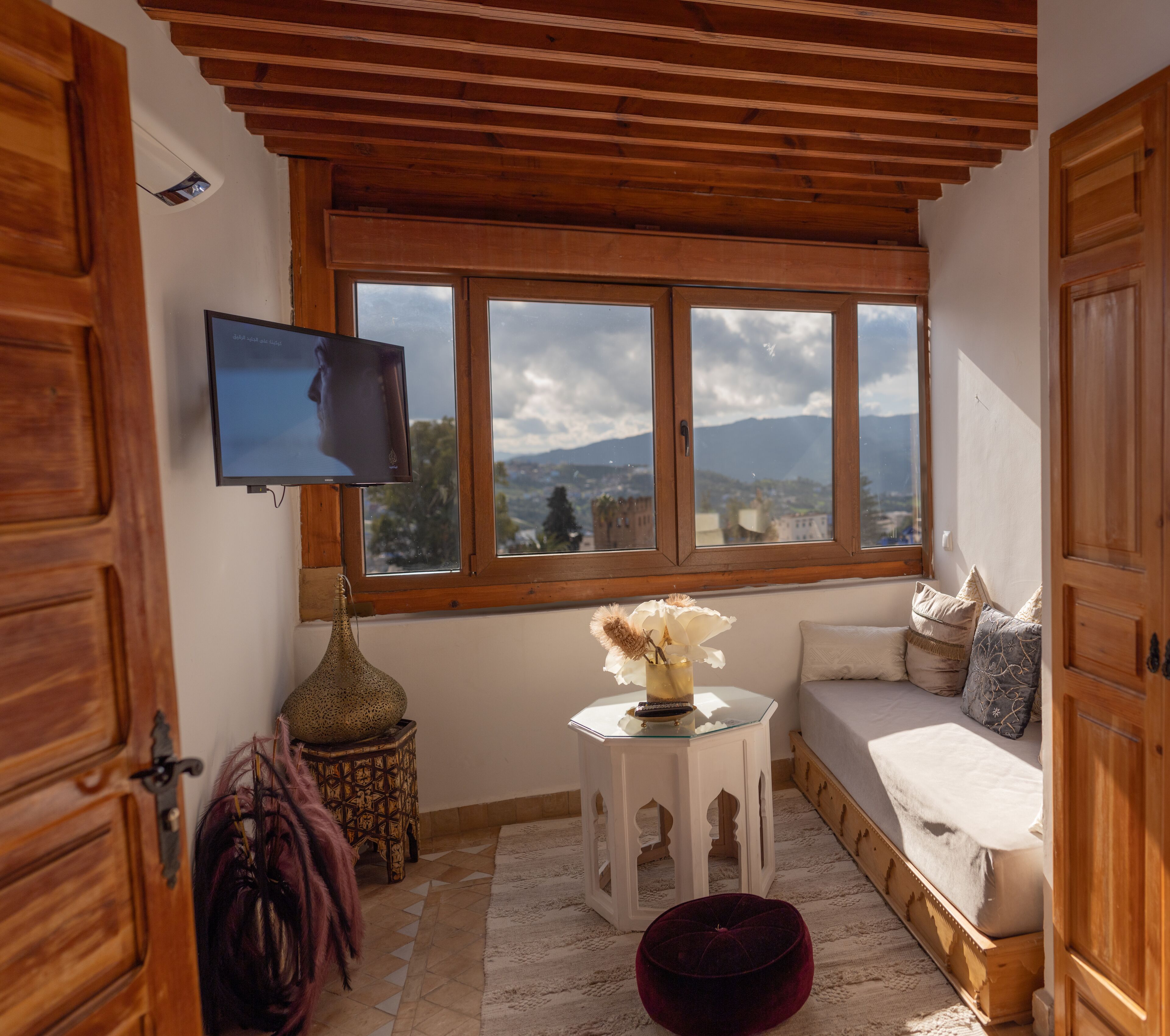 Une pièce chaleureuse avec un plafond en bois, une grande fenêtre donnant sur les montagnes, une télévision, une petite table avec un vase et un banc rembourré.