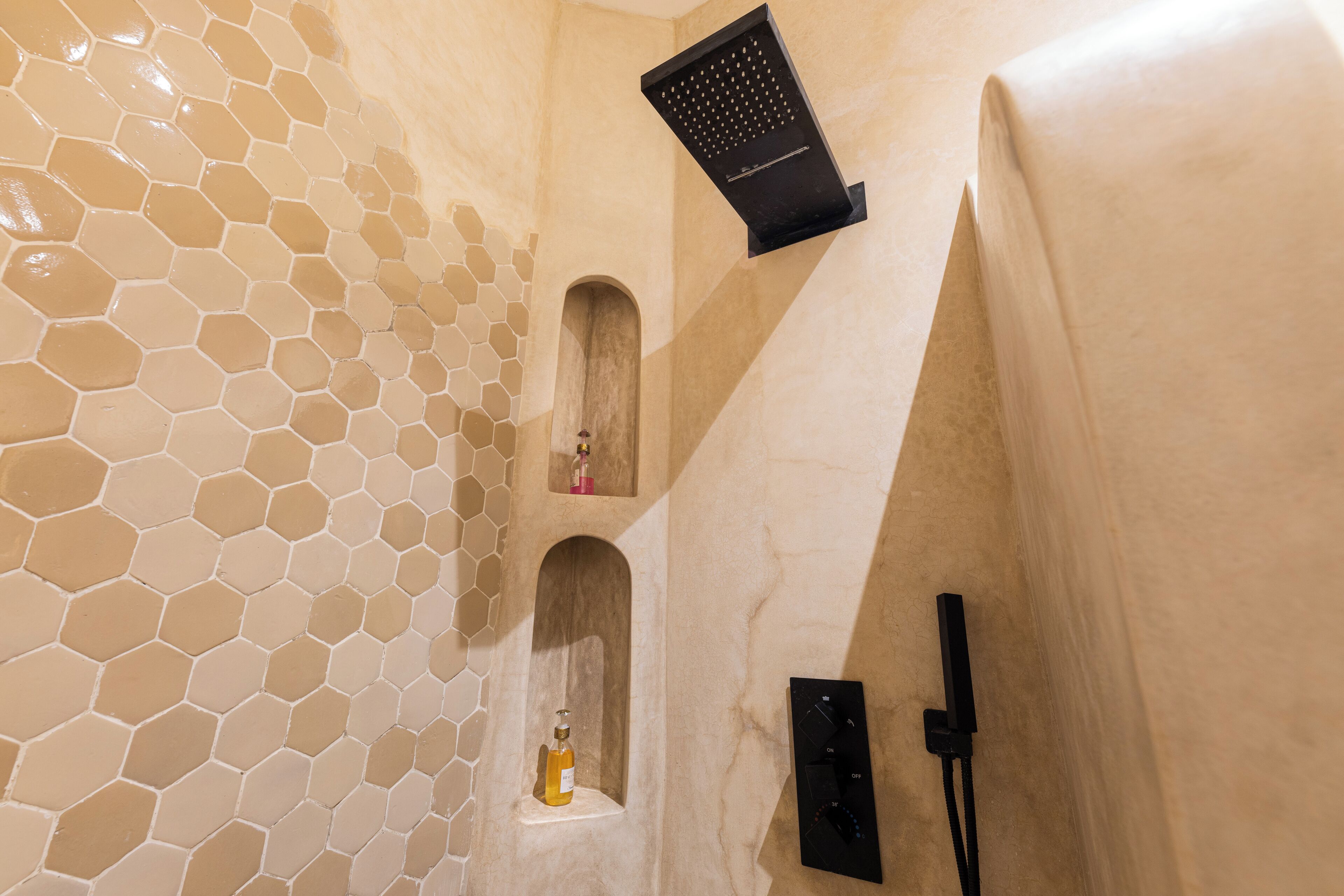 A bathroom with hexagonal tile walls, a showerhead, and a soap dispenser.