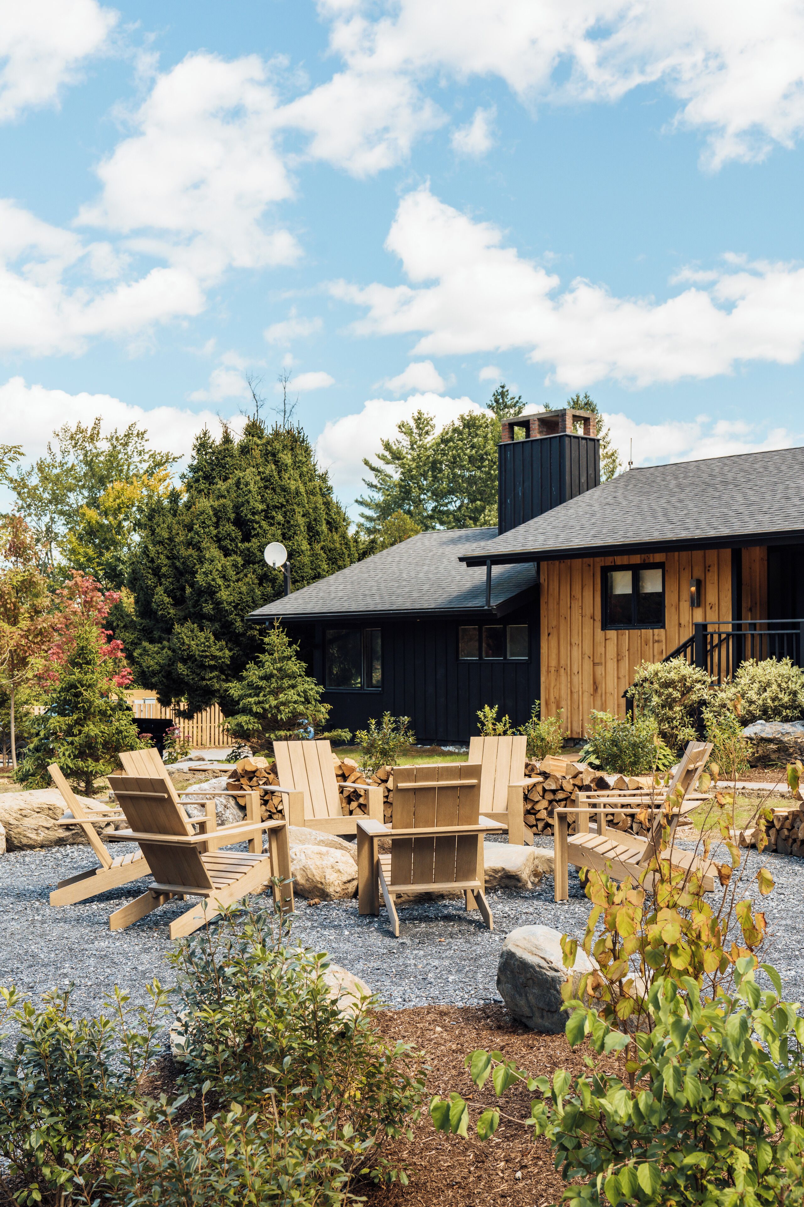 A modern house with a wooden exterior, a black roof, and a gravel patio with wooden chairs and a fire pit.