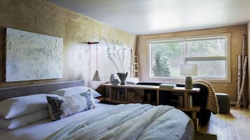 A bedroom with a bed, a desk, a window, and artwork on the wall.
