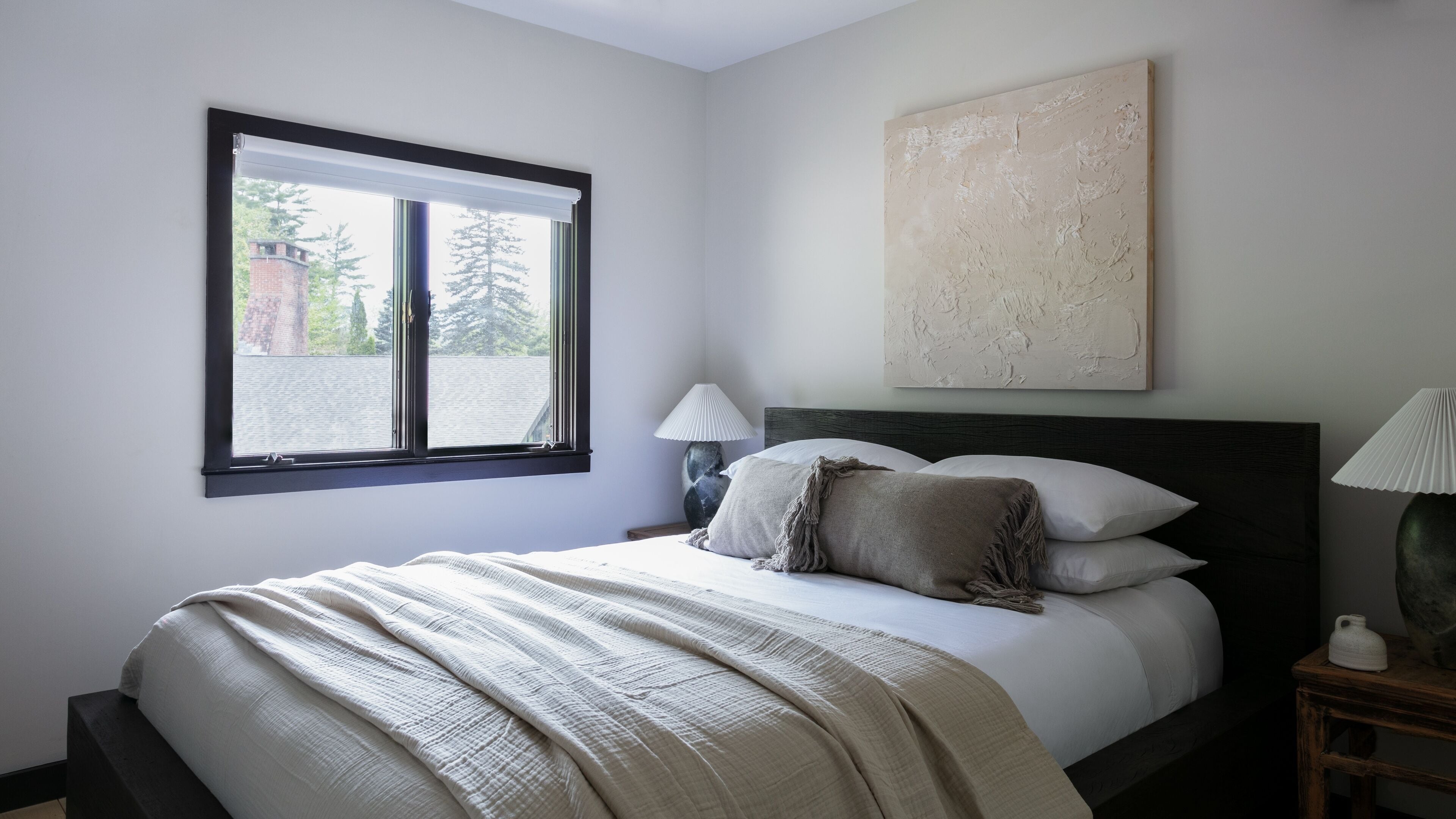 A bedroom with a bed, a nightstand, a lamp, a window, and a painting on the wall.