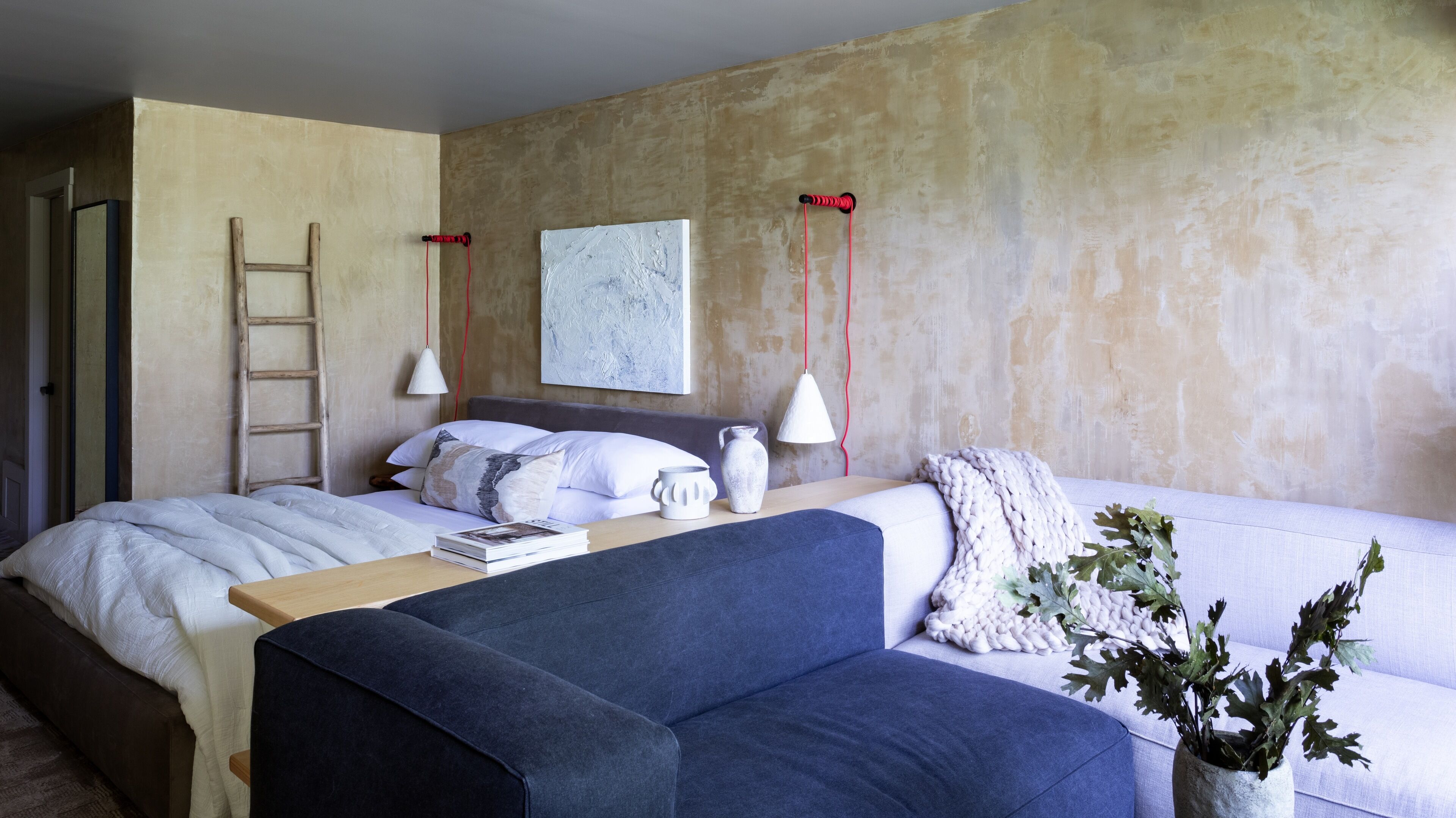 A bedroom with a bed, a blue sofa, a wooden nightstand, a white vase, and a painting on the wall.