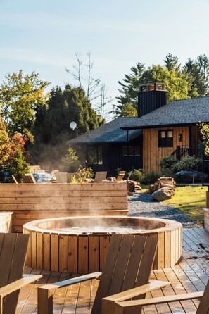 En træterrasse med et spabad omgivet af Adirondack-stole, en træhytte med skorsten og en skov i baggrunden.