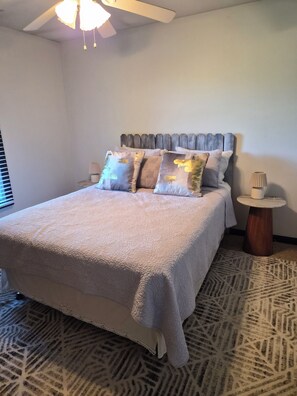 Una cama bien hecha con un edredón texturizado, cabecera de madera y almohadas decorativas. Junto a la cama, una mesita redonda con una lámpara.