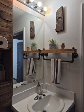 Un baño con lavamanos blanco, un espejo, toallero y estante con plantas en macetas.