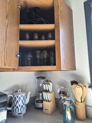 A kitchen with a wooden cabinet containing wine glasses, a blender, a kettle, and a knife block.