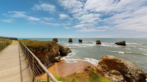 A coastal walkway with a wooden boardwalk and metal railing, leading to a rocky shoreline with waves crashing against large boulders.