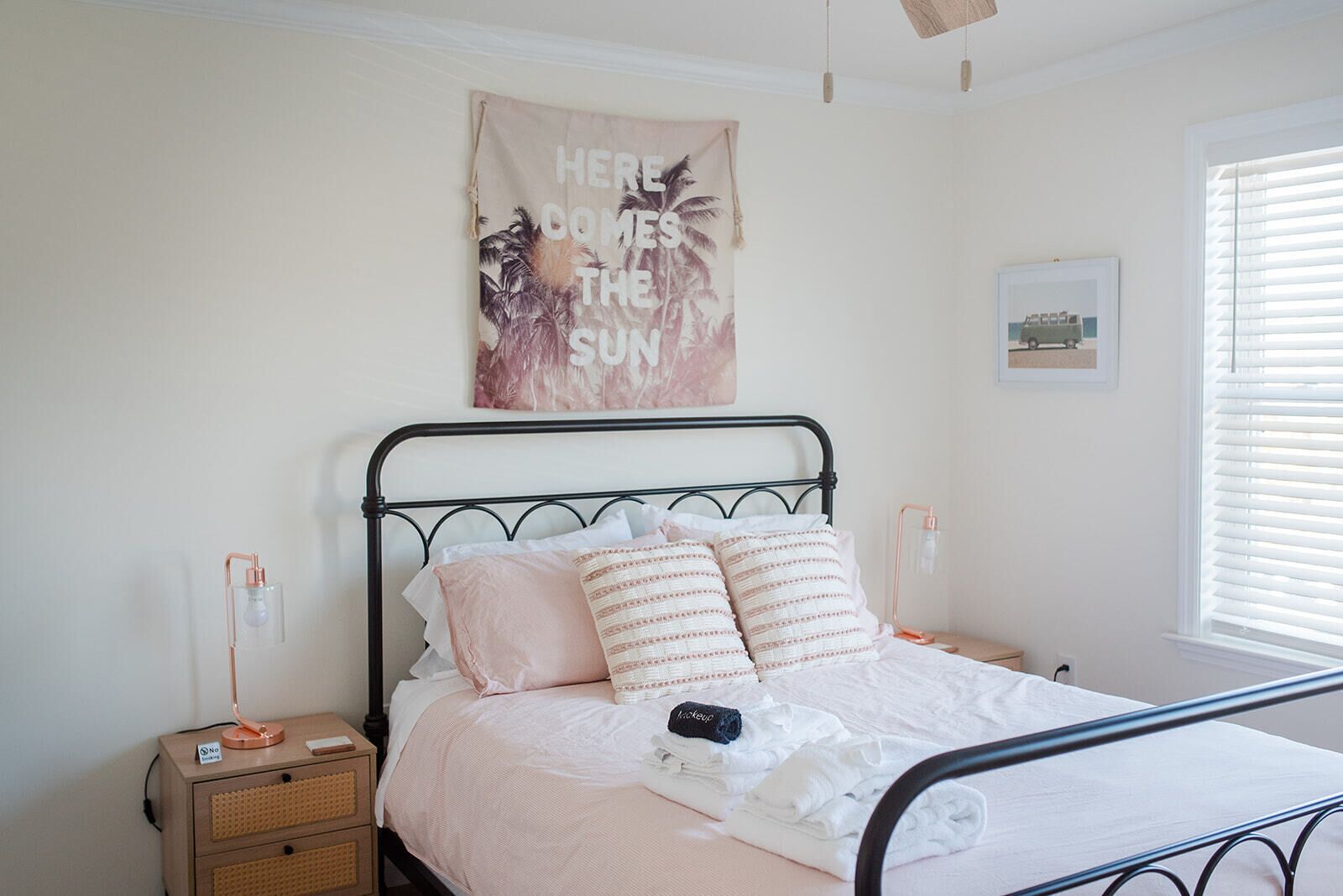 A neatly made bed with a pink and white bedding, a small bedside table with a lamp, and a wall hanging with a tropical design.