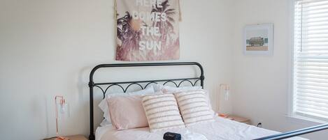 A neatly made bed with a pink and white bedding, a small bedside table with a lamp, and a wall hanging with a tropical design.