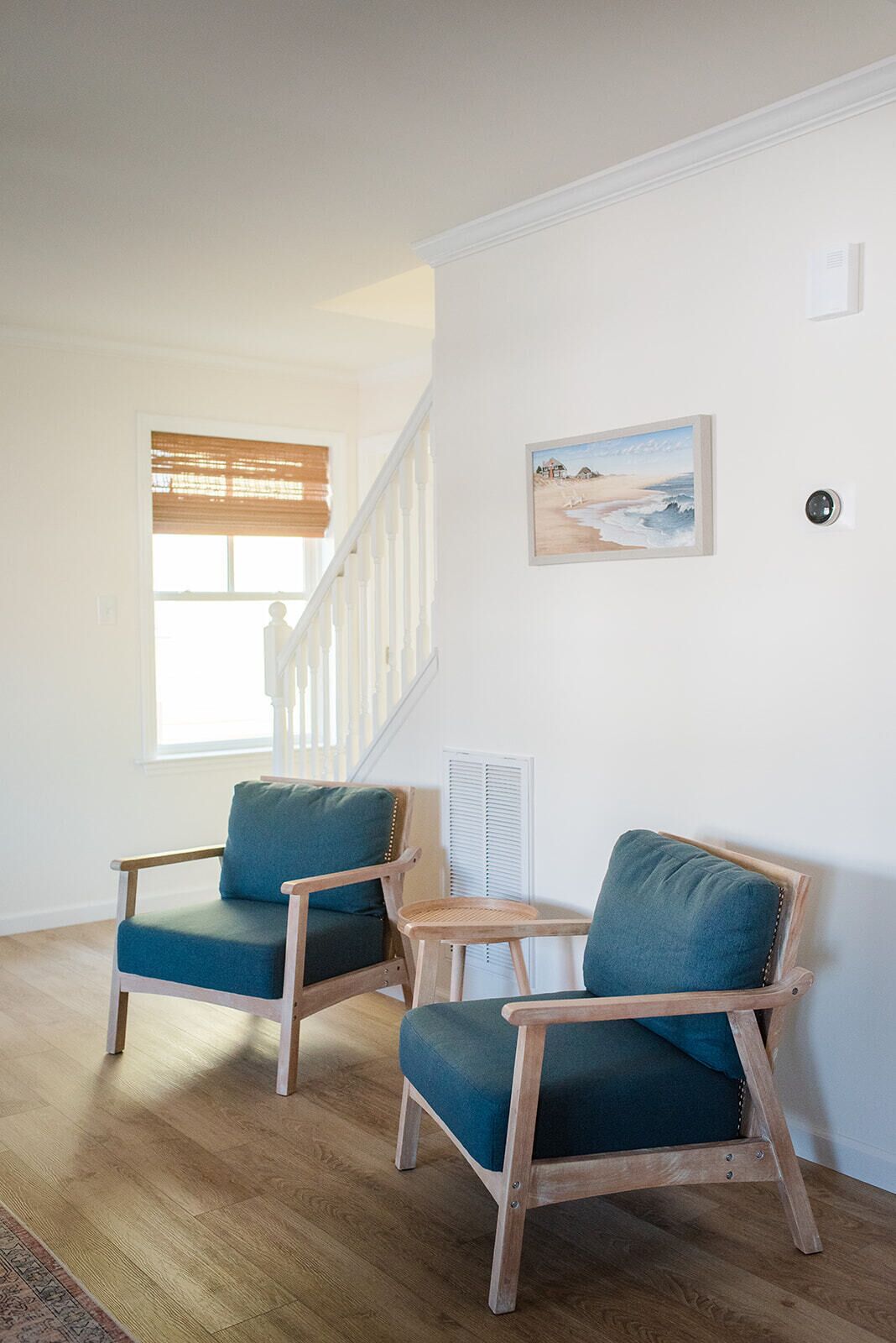 Une pièce avec deux chaises en bois dotées de coussins turquoise, un radiateur blanc, un tableau représentant une plage, encadré, et un escalier avec des rampes blanches.