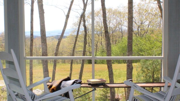 A porch with two rocking chairs, a table, and a view of a forest and mountains. - Rustic Chic Maine Cabin (Sedgwick)