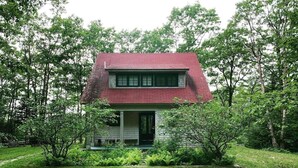 A two-story house with a red roof, surrounded by lush greenery and large rocks in the foreground. - Rustic Chic Maine Cabin (Sedgwick)