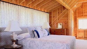 A bedroom with a wooden ceiling, a bed with a floral bedspread, two bedside lamps, and a dresser with a mirror. - Rustic Chic Maine Cabin (Sedgwick)