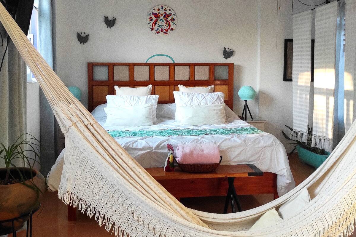 A bedroom with a hammock, a bed with white bedding, a ceiling fan, and a decorative wall hanging.