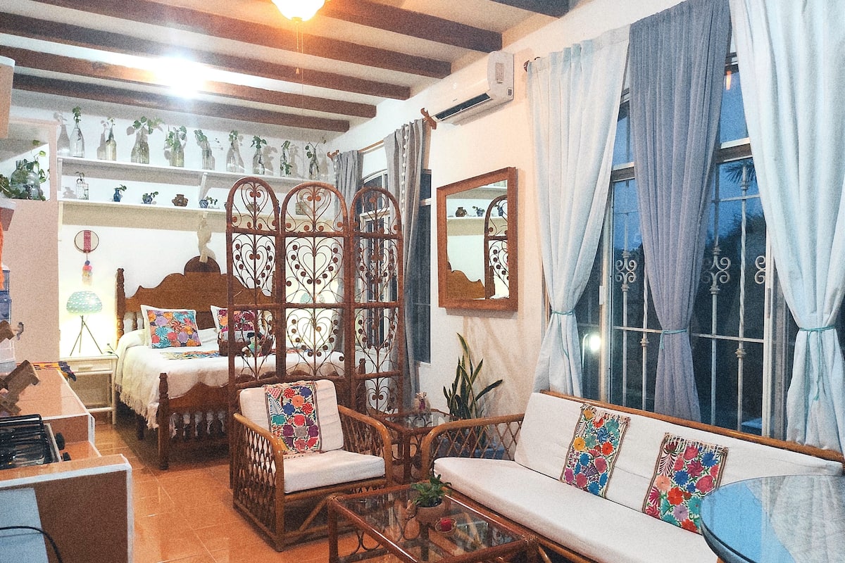A cozy room with a bed, a sofa, and a dining table. There is a decorative screen, a mirror, and a window with curtains.