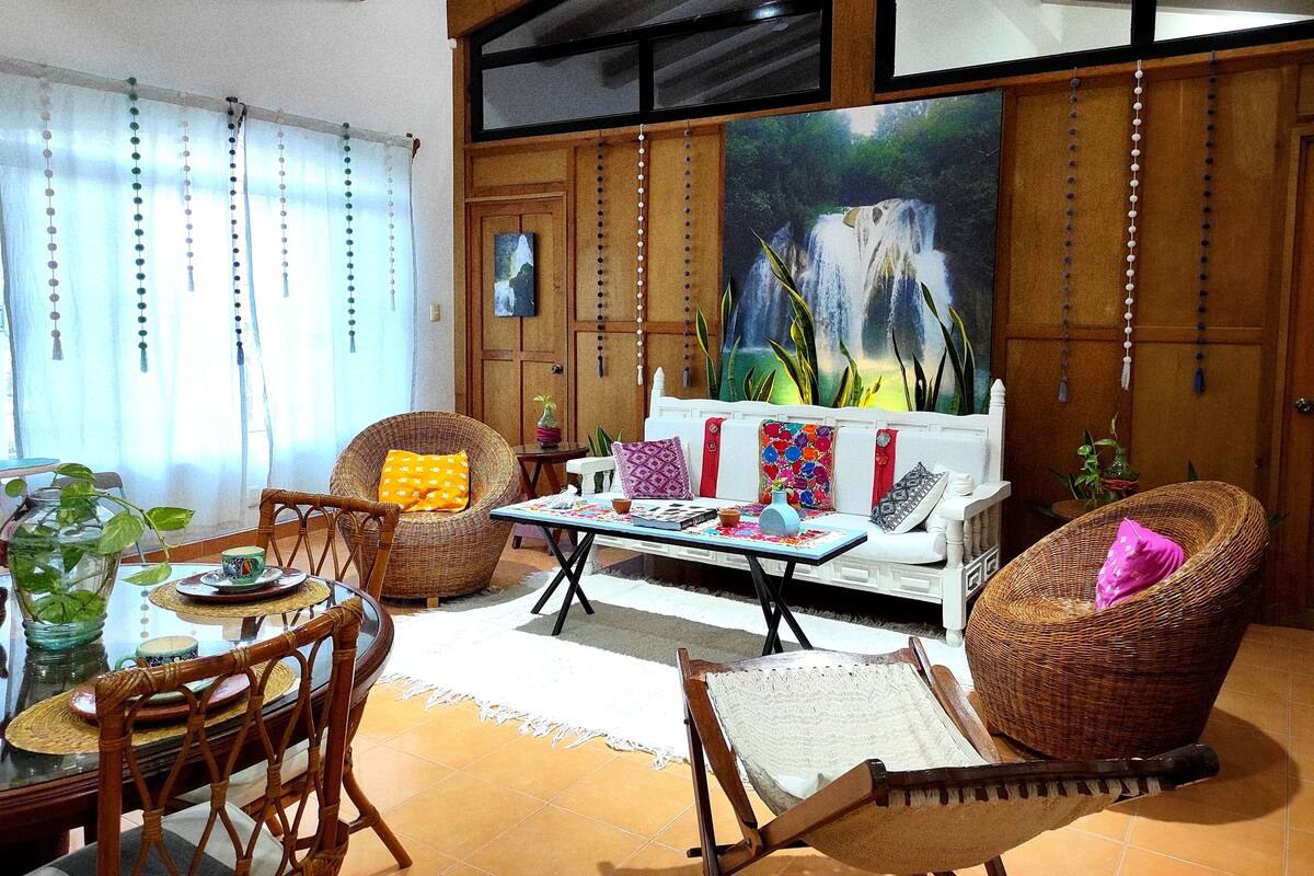 A cozy living room with wicker furniture, a dining table with a vase of flowers, and a large painting of a waterfall.