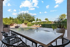 Een terras buiten met een rechthoekige tafel en stoelen, een kleine waterval en parasols voor schaduw.