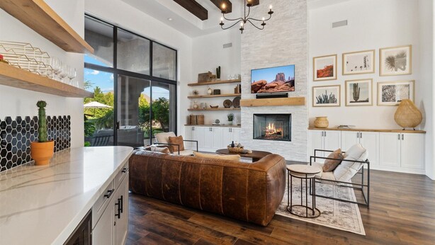 A spacious living area with a fireplace, large windows, and a modern kitchen with wooden beams and a stone wall. - Scottsdale Paradise- POOL, Spa, Pickleball/Basketball, Fire Pit & Great HIKING! (Scottsdale)