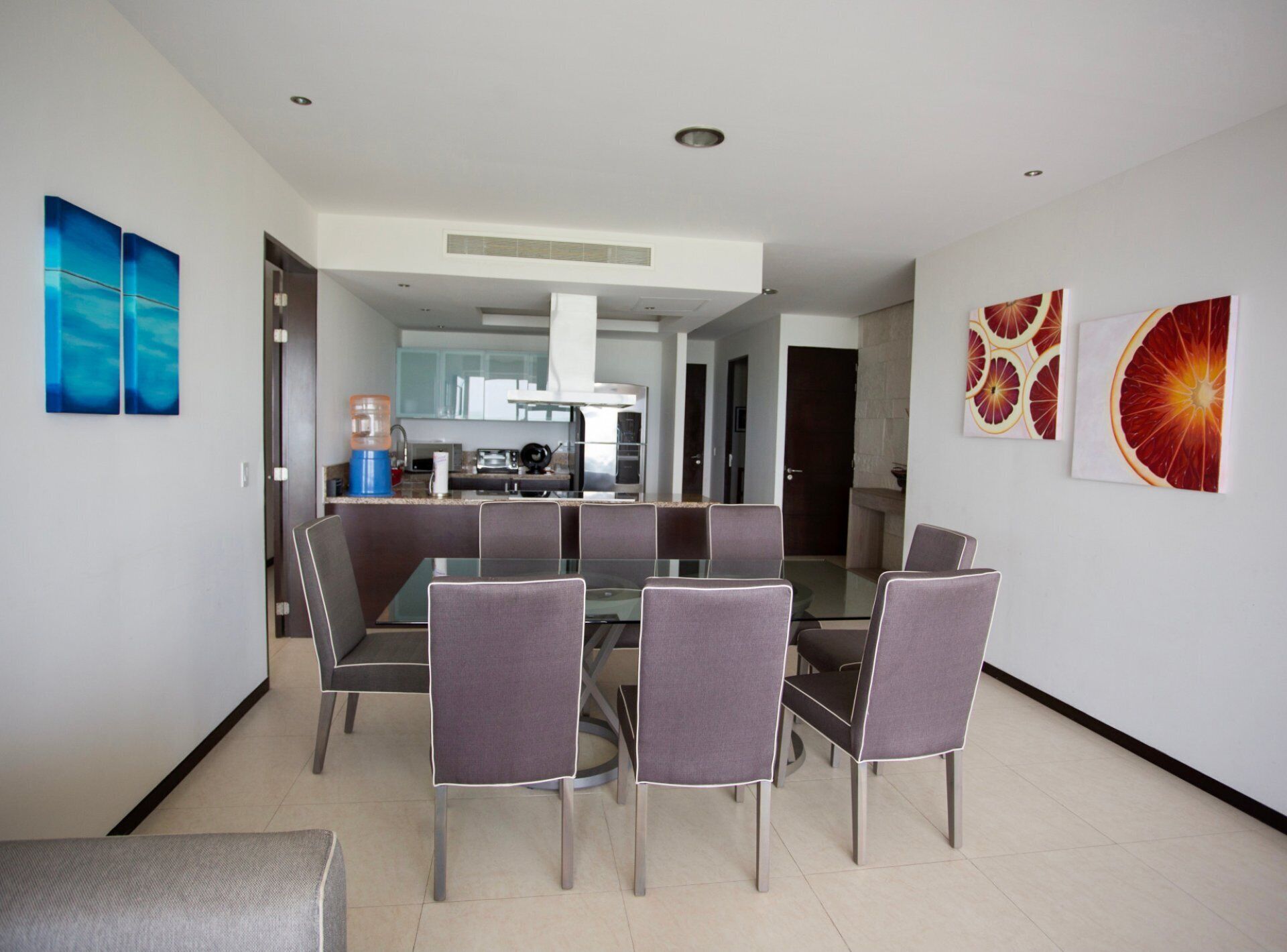 A modern dining area with a glass table and six chairs, a kitchen in the background, and two large fruit-themed paintings on the walls.