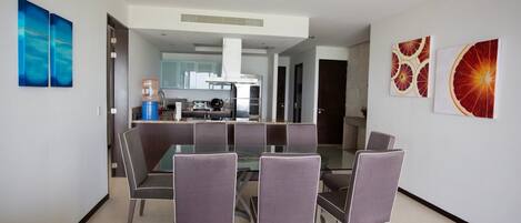 Un comedor moderno con una mesa de vidrio y seis sillas, una cocina al fondo, y dos grandes cuadros con motivos de frutas en las paredes.