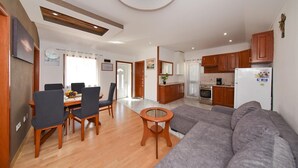 Ein modernes Wohnzimmer mit grauem Sofa, Esstisch aus Holz und Stühlen. Es gibt eine Küchenzeile mit Holzschränken und einem Kühlschrank.