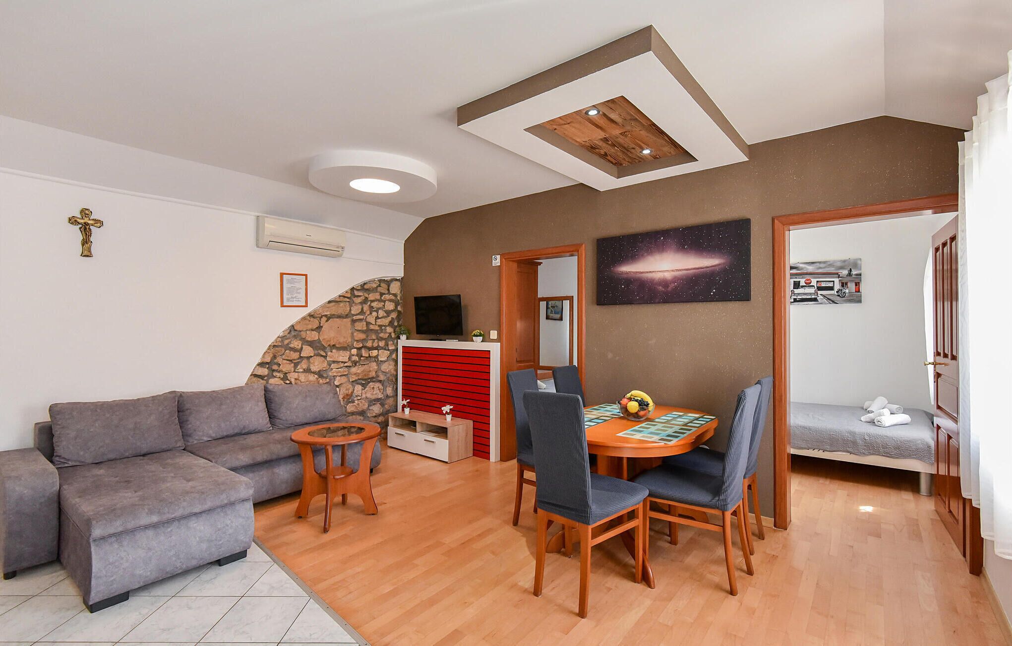 A living room with a grey sofa, a wooden dining table with chairs, a small round table with a plant, and a stone wall.