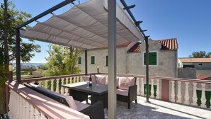 Ein überdachter Außenbereich mit Tisch und Stühlen, Blick auf ein Steinhaus mit einem Ziegeldach und einem klaren, blauen Himmel.