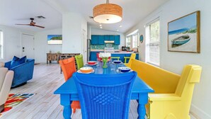 A bright dining area with a blue table and yellow and orange chairs, a ceiling fan, and a kitchen in the background. - Island Adventures Await at the Pet Friendly “Boathouse SGI” (Saint George Island)