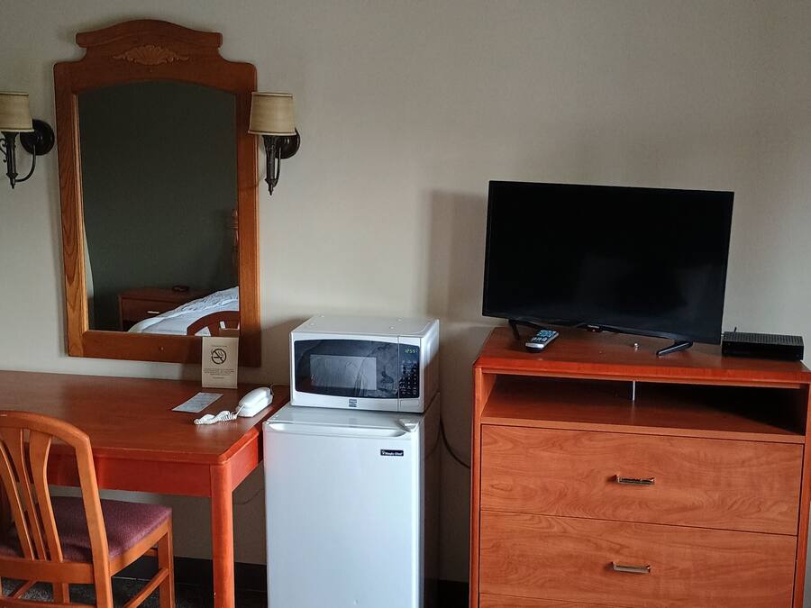 Habitación de hotel con un escritorio de madera, un refrigerador blanco, un microondas, un televisor sobre un mueble de madera, un espejo con apliques de pared y una cama.
