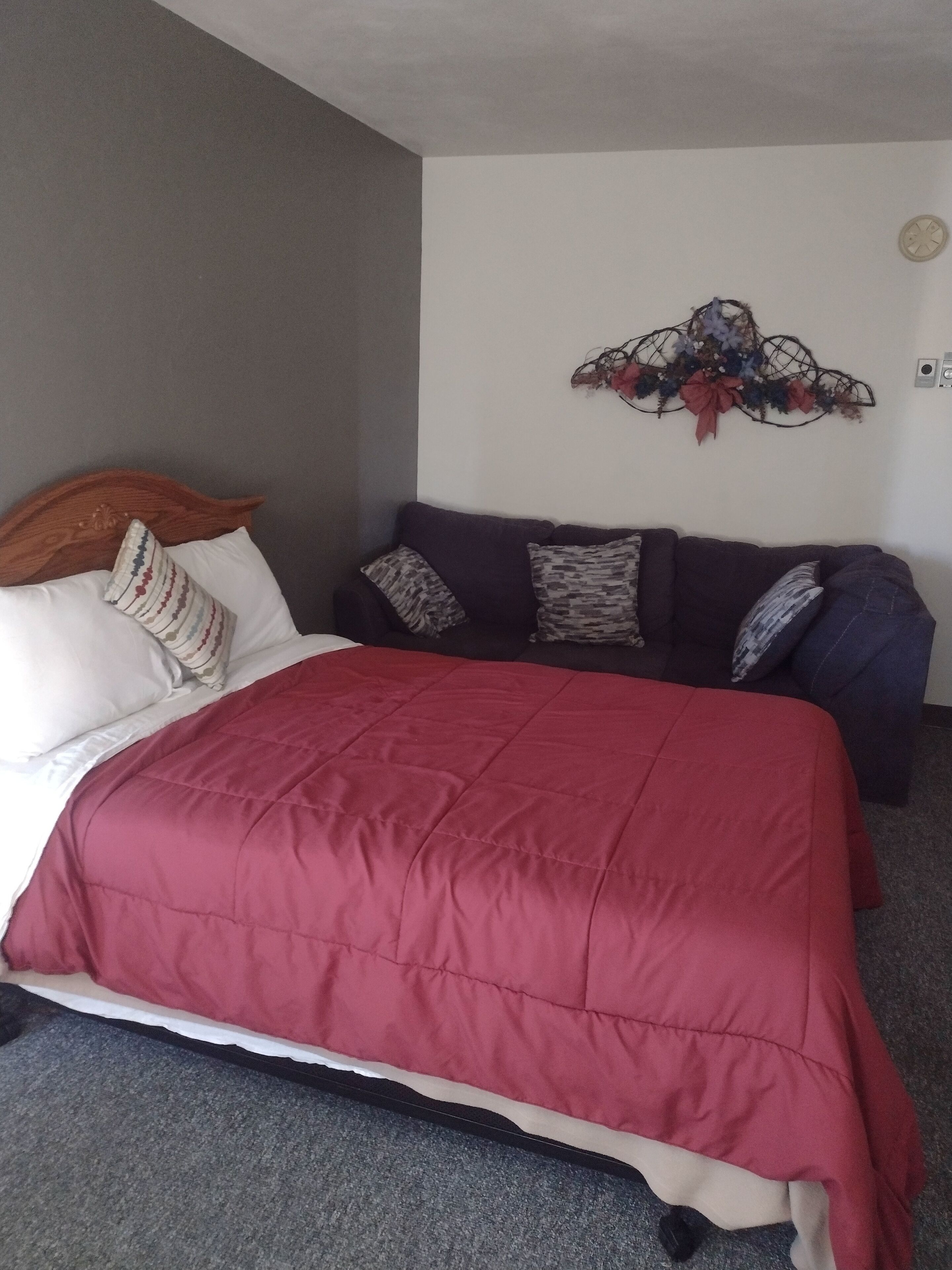 A bedroom with a bed, a dark-colored sofa, a decorative wall piece, and a carpeted floor.
