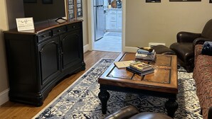 A living room with a dark wooden cabinet, a television, a coffee table with books, a brown leather sofa, and a patterned rug.
