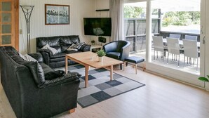 Ett vardagsrum med en soffa, fåtöljer, ett soffbord och en tv. Det finns ett stort fönster med utsikt över omgivningen.
