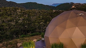 A geodesic dome structure with a stone base and a grassy foreground, set against a backdrop of a forested hillside and a clear sky. - Kairos Glamping - Rancho Queimado - SC (Rancho Queimado)