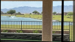 Une vue depuis un balcon orné d’une colonne en pierre, avec une fontaine et un lac en contrebas, des montagnes se dessinant à l’horizon.
