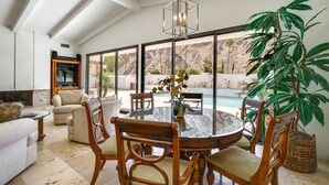 A spacious living room with a round glass table, wooden chairs, a sofa, and a large window overlooking a pool. - Private Pool & Spa: Indian Wells Home! (Indian Wells)