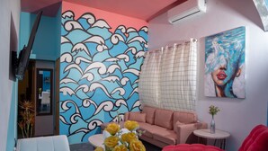 Ein modernes Wohnzimmer mit blauen und pinkfarbenen Akzenten, einer Wand mit großem Wellenmuster, einem pinken Sofa, einem roten Sessel und einem runden Tisch mit einer Vase voller gelber Blumen.