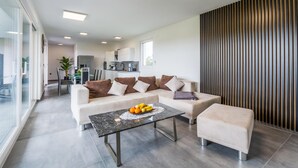 Ein modernes Wohnzimmer mit Sofa, Couchtisch und Pouf. Im Hintergrund befindet sich eine Küche mit weißen Schränken und einem Essbereich.