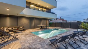 Ein moderner Poolbereich im Freien mit Liegestuhl und Tisch, einer überdachten Sitzgruppe und einem zweigeschossigen Gebäude mit großen Fenstern.