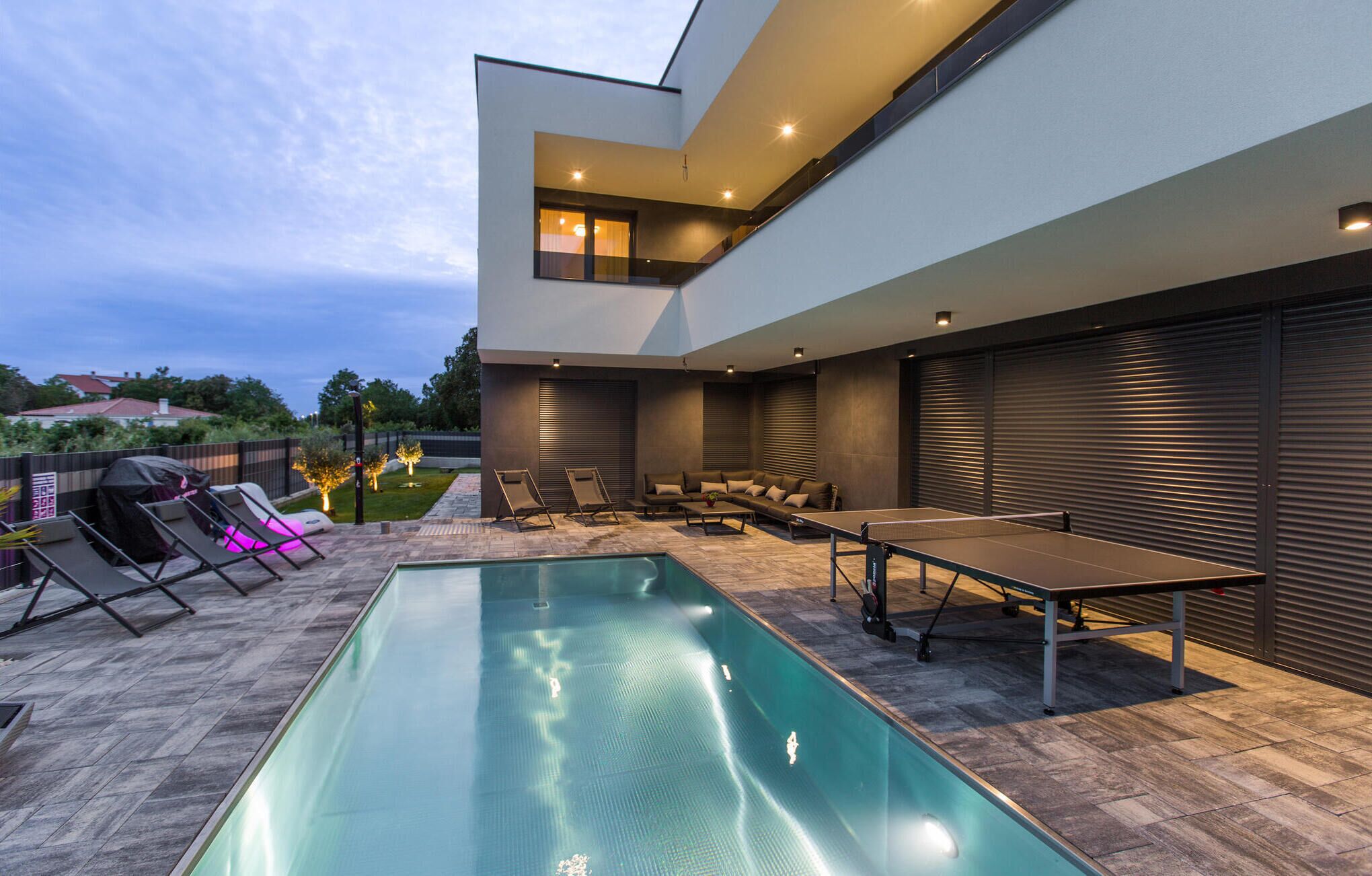 Ett modernt utomhusområde med pool, ett bord för bordtennis, solstolar och en överbyggd sittplats.