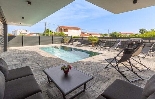 Área de piscina no terraço, com mesa, cadeiras, churrasqueira e vista para edifícios ao redor.