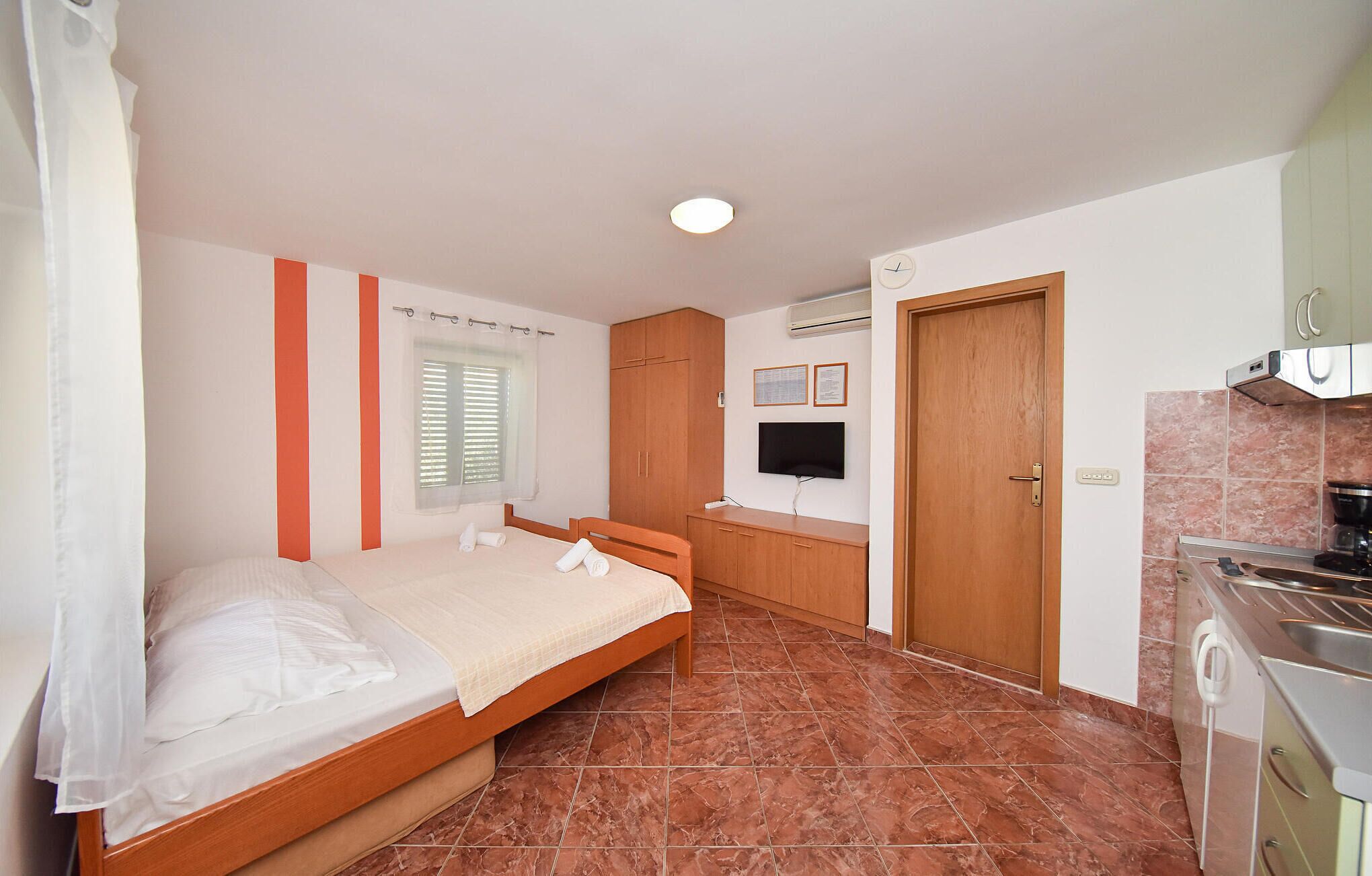 Ein Schlafzimmer mit einem Bett, einem Fenster mit Jalousien, einem Holzschrank, einem Fernseher und einer Küchenzeile mit Herd.