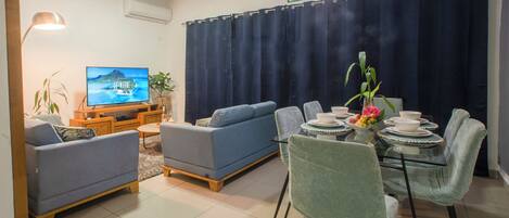 Un salon comprenant un canapé, un téléviseur, une table à manger avec des chaises et un rideau.