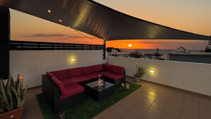 Area esterna con divano e tavolo, una catena di luci e vista sul tramonto sull'oceano.