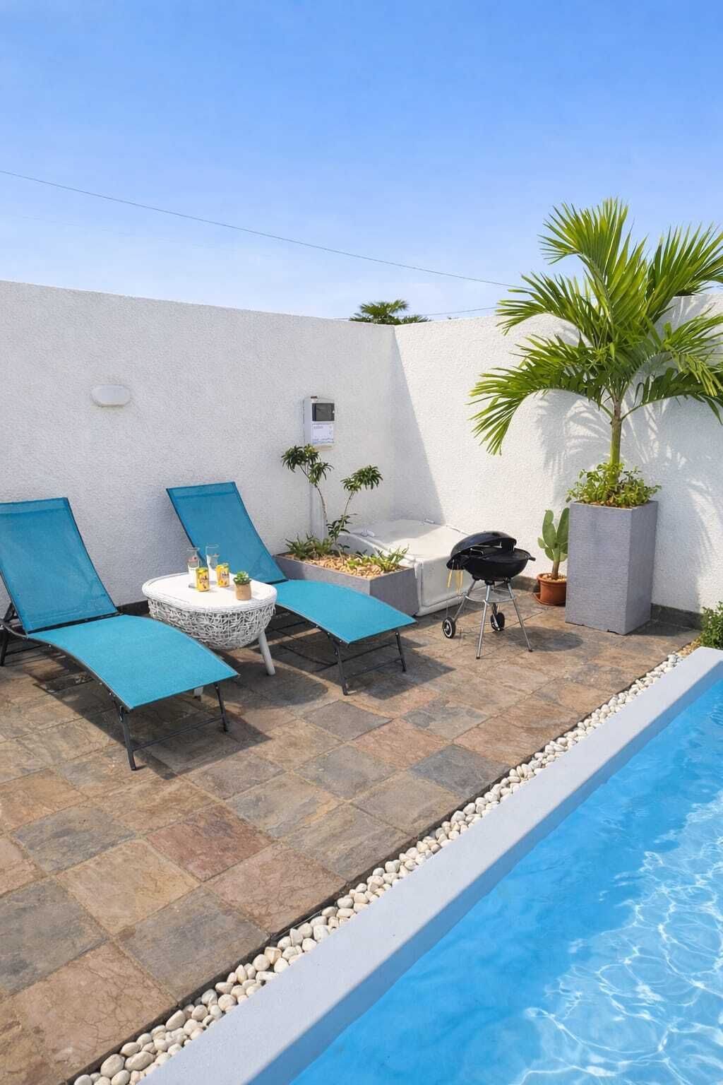 Ein Poolbereich mit zwei Liegestühlen, einem kleinen runden Tisch mit Getränken, einem Grill und einem Blumentopf.