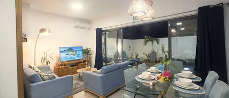 Una sala de estar con sofá, televisor, mesa de comedor con sillas y un balcón con plantas.