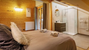 A bedroom with a wooden wall, a bed with rolled towels, a bedside table, a mirror, a bathroom with a shower, and a radiator.