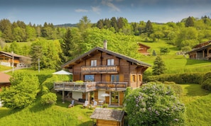 A wooden chalet with a balcony, surrounded by lush greenery and a mountainous backdrop.